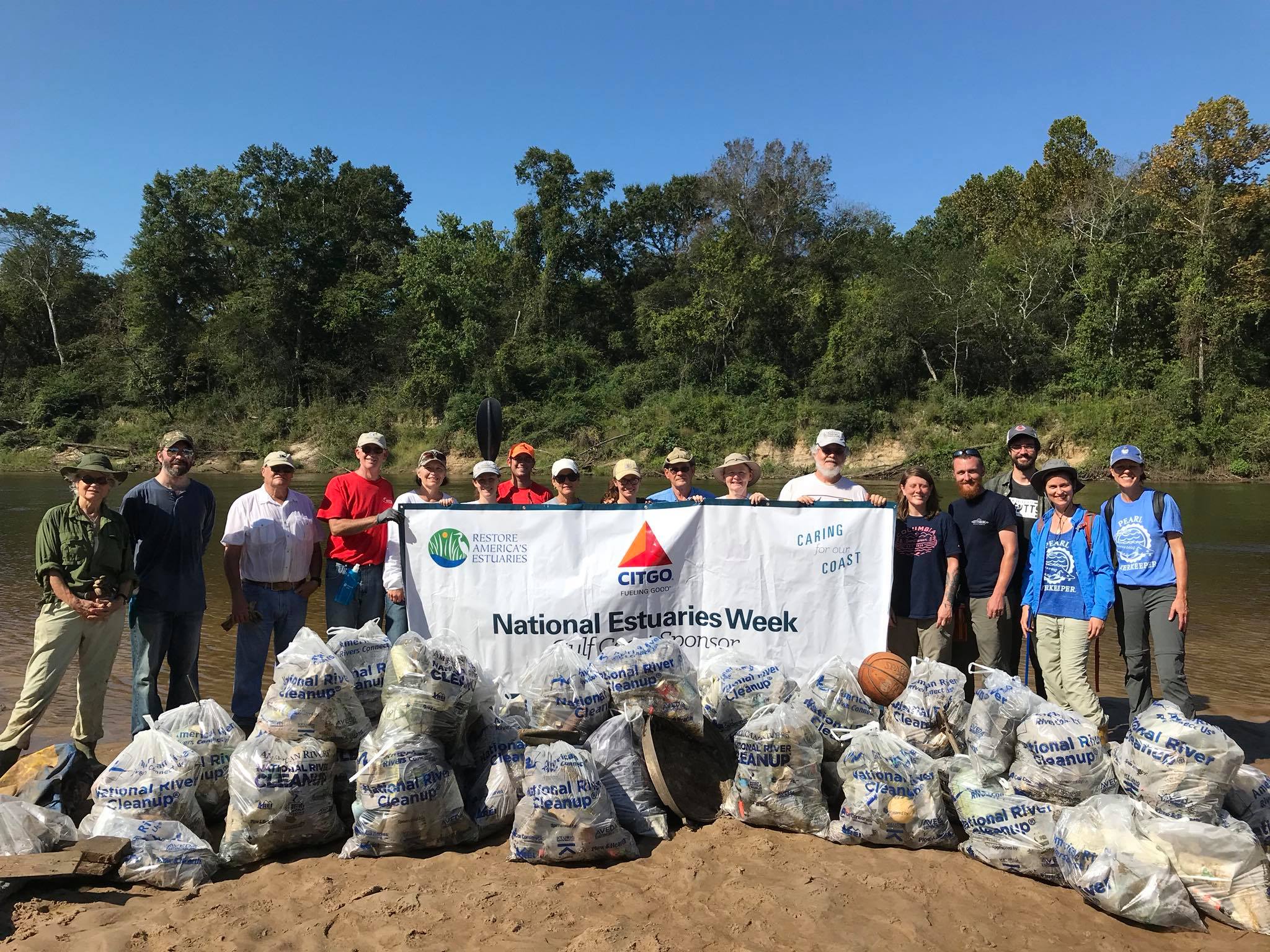 Group of 17 volunteers posting with a banner and many bags of trash at a river clean up project.