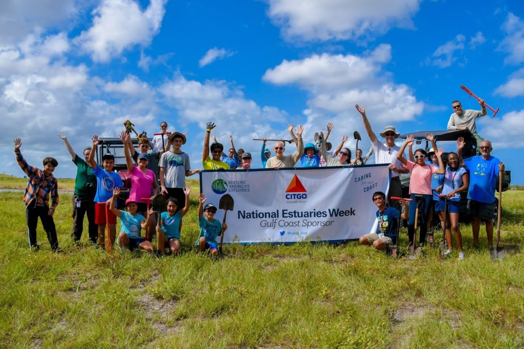 Group of 28 people standing in coastal area holding up a banner for National Estuaries Week